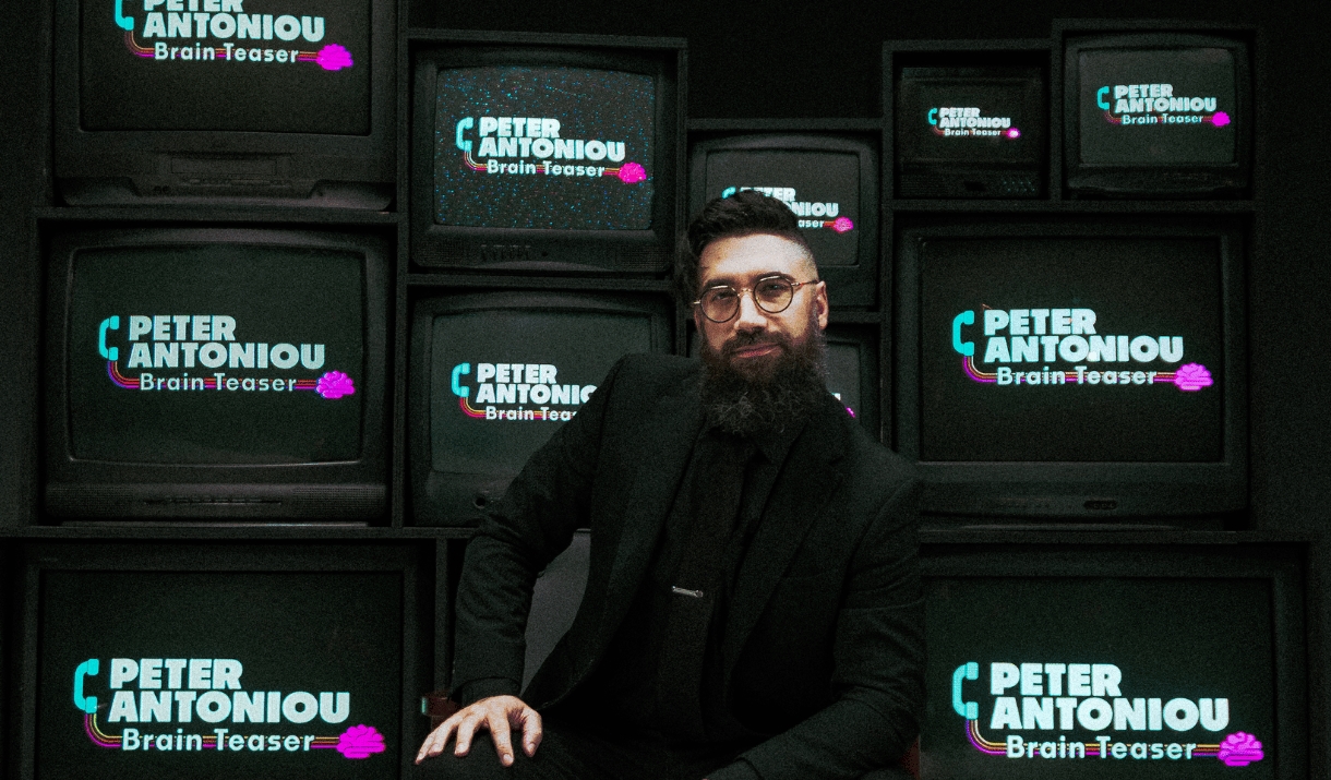 A man in round glasses and a black suit poses in front of TVs showing the Brain Teaser Logo.