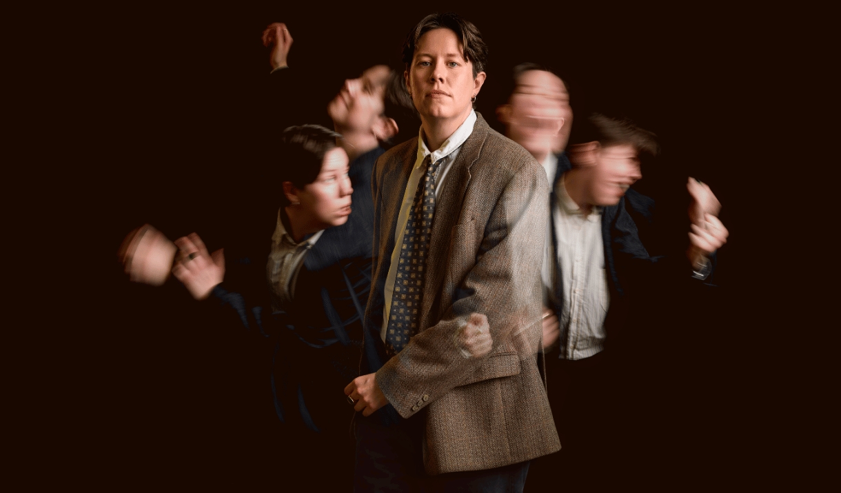 Chloe Petts in a tweed blazer stands still while blurred versions of thermselves move around them in the background.