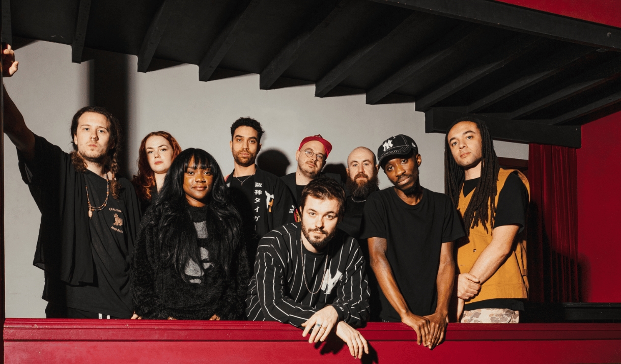 A nine-piece band poses behind a red balcony rail beneath dark wooden beams.
