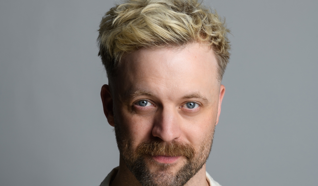 Headshot of Thomas Green with bleached blonde hair and a beard, looking at the camera against a grey background.