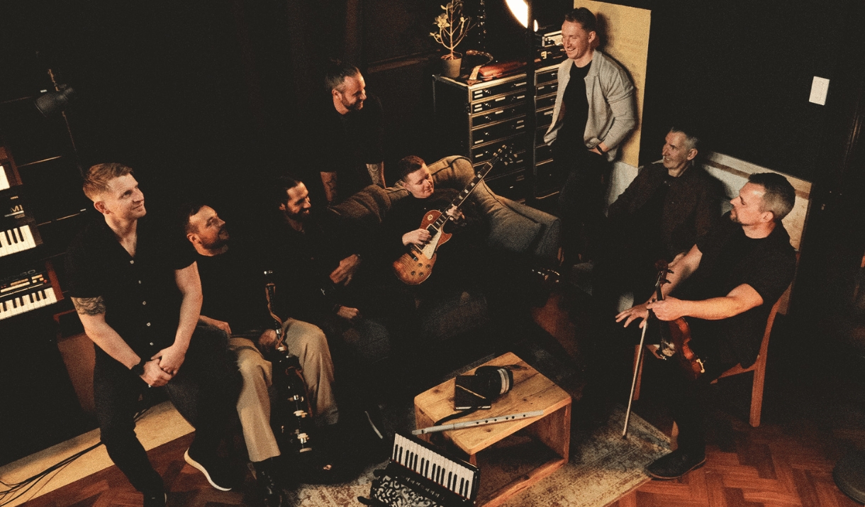 Photo of 8 band members sitting in a relaxed studio. Some of them are holding instruments including  a guitar and a violin.