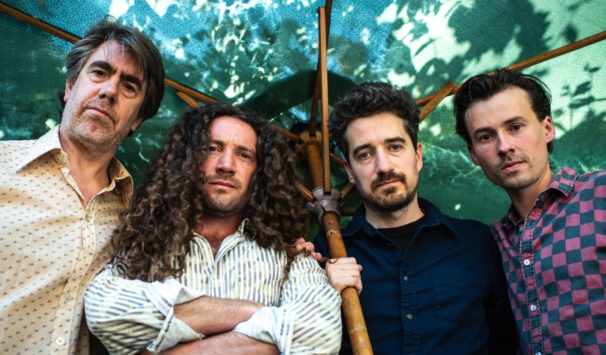 Four-piece band posed together under a large parasol against a green leafy backdrop.