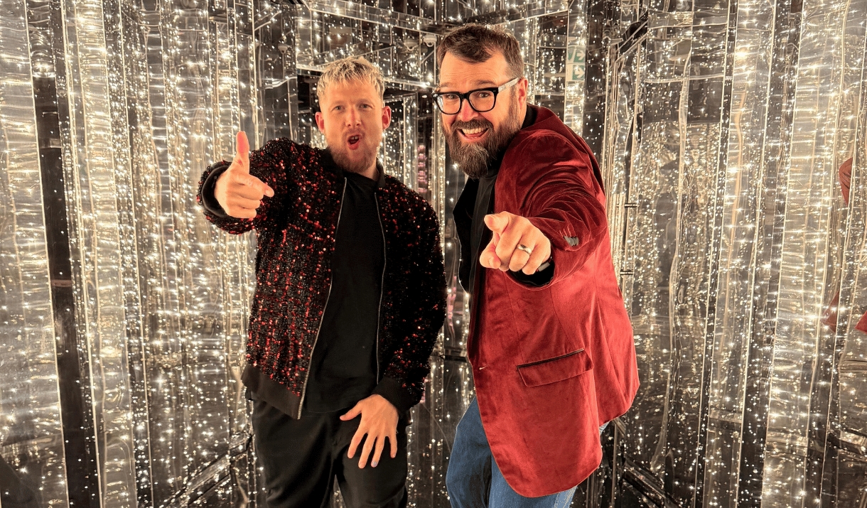 Two grinning men point at the camera among shimmering fairy lights.