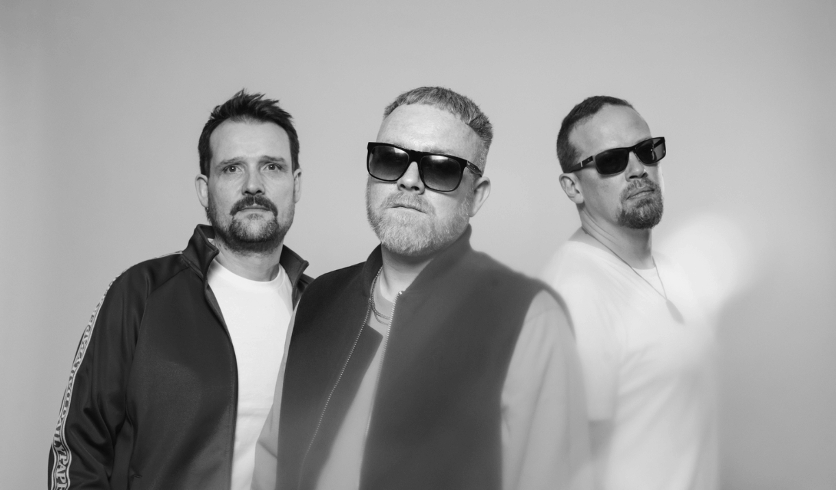 Black and white image of three men against a grey background. Two of them wearing sunglasses, all looking serious.