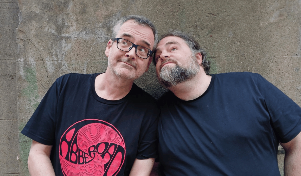 Two men in dark T-shirts leaning their heads together and pulling funny faces against a weathered stone wall.