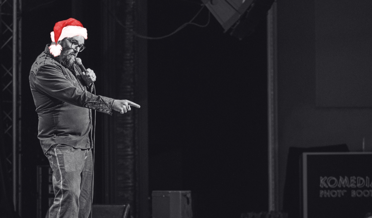 A comedian in red Santa hat points at the audience on stage in a black-and-white photo.