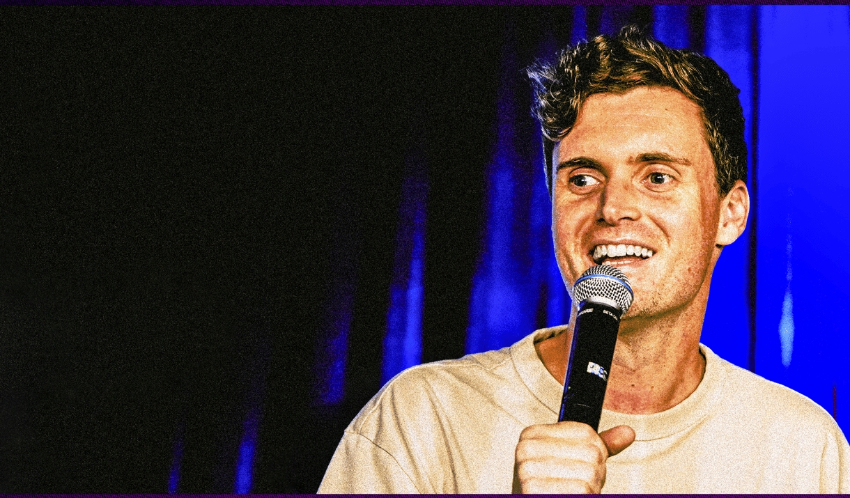 A smiling comedian holds a microphone against a blue stage curtain.