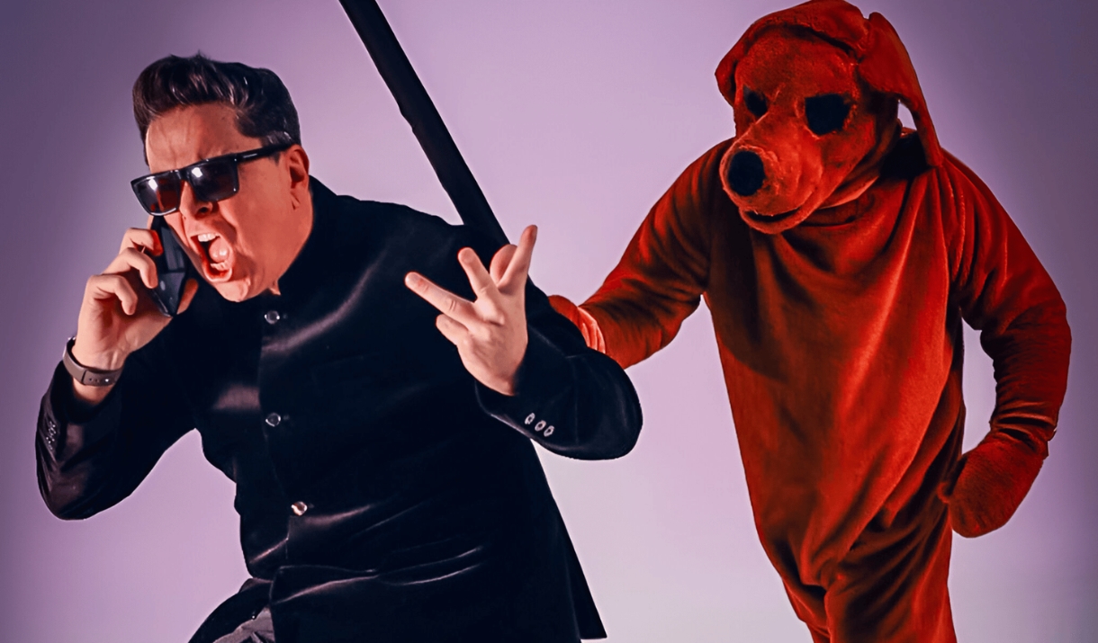 A man in a black suit and sunglasses poses dramatically beside a person in a red dog costume.