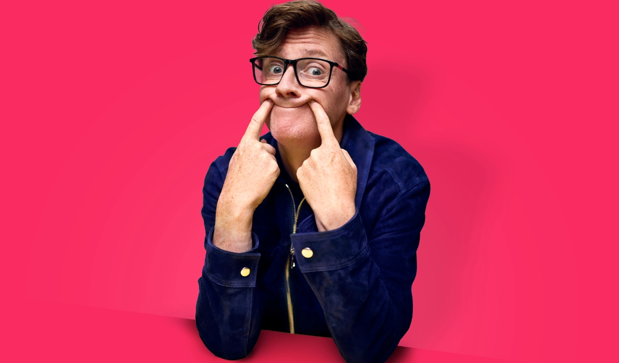 A comedian in glasses and a blue jacket pulls a silly face against a bright pink background.