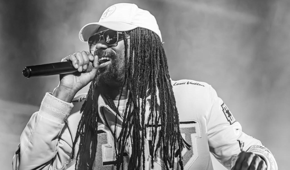 Black and white photo of a MC with long dreadlocks performing into a microphone, wearing sunglasses and a cap.
