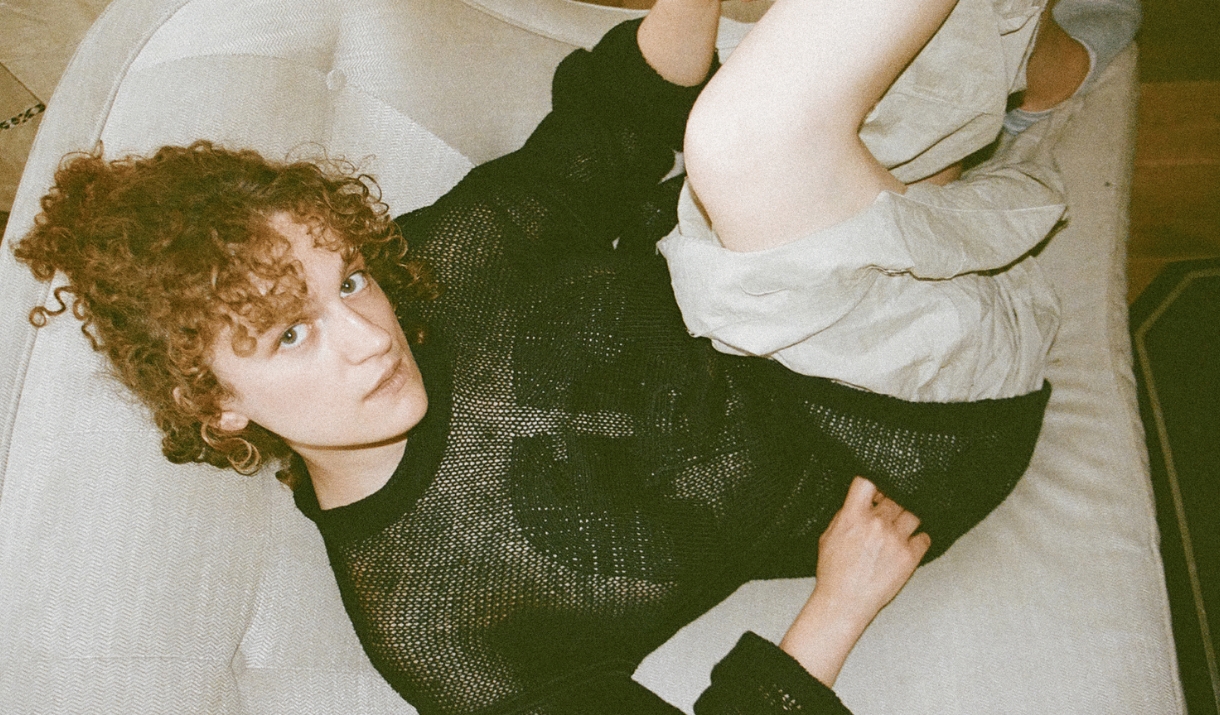 A person with curly red hair in a black mesh top lies on a white sofa, looking up at the camera.