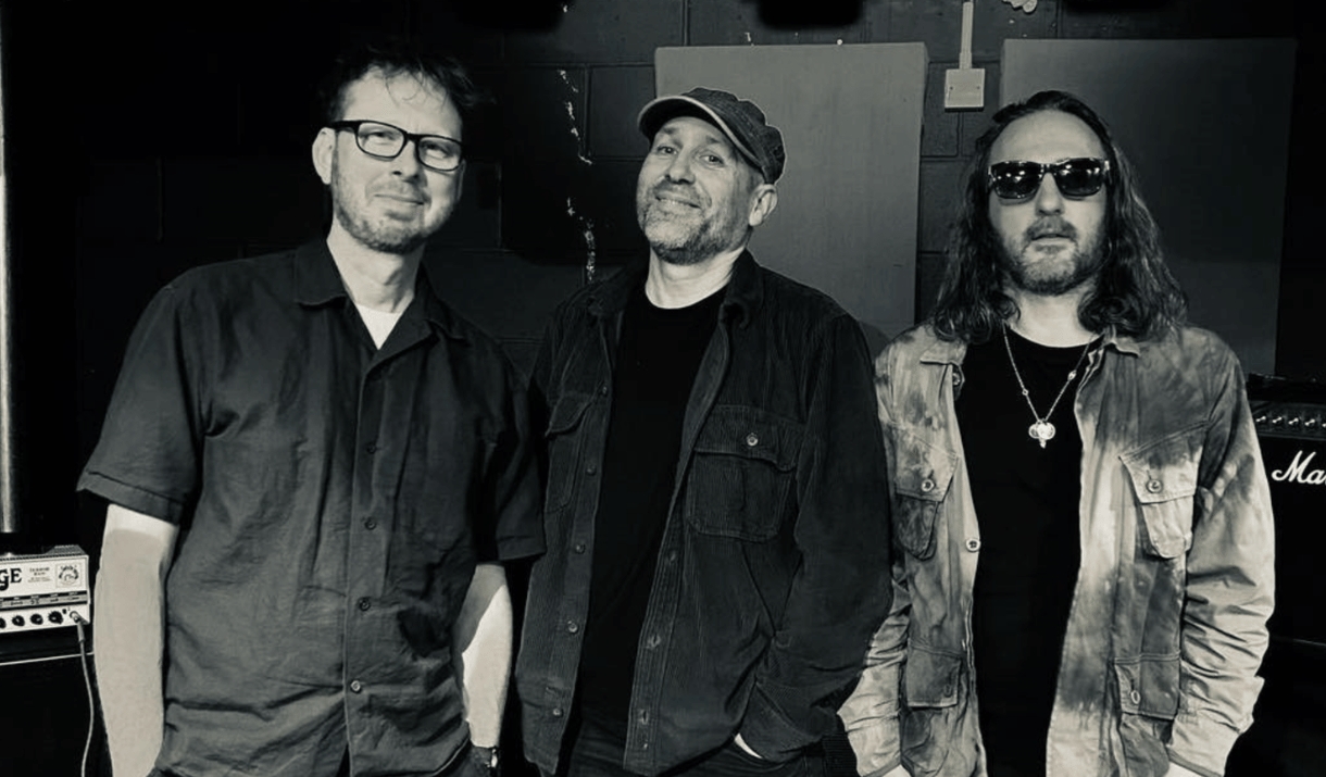Photo of three male band members against a dark studio background. One wearing glasses, one wearing a cap and one wearing sunglasses. All smirking.