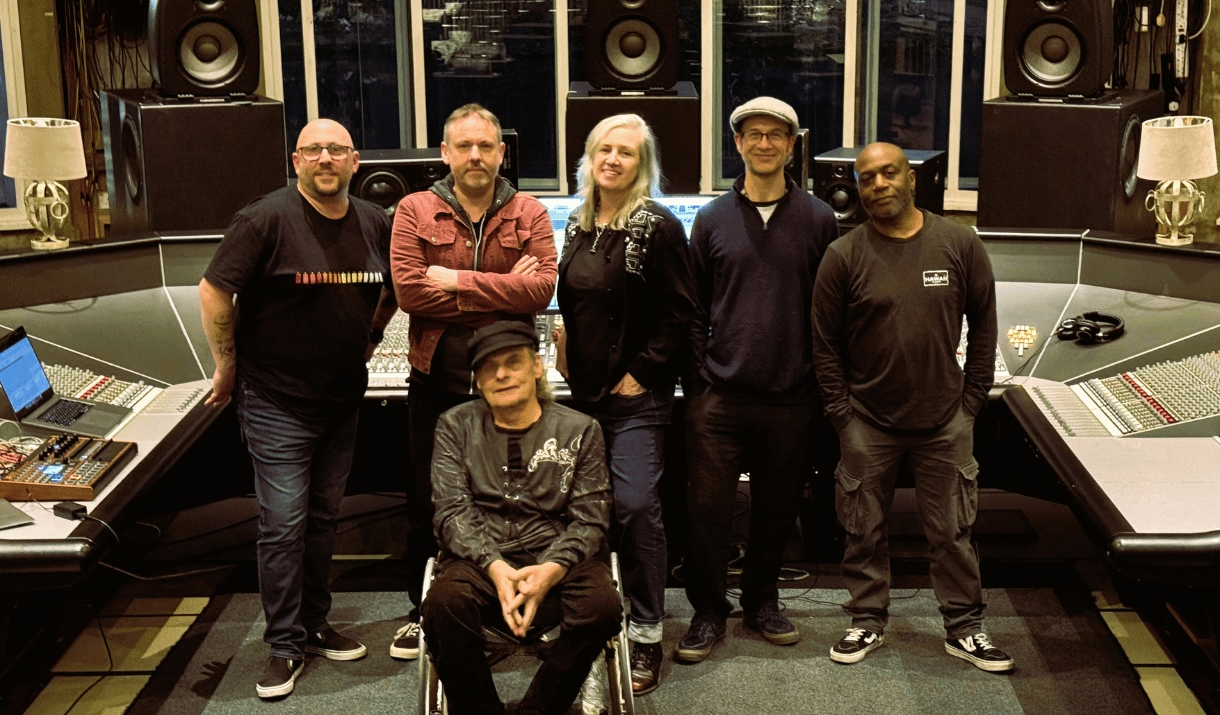 Six-piece band posing together in a recording studio, in front of a large mixing desk and studio monitors.