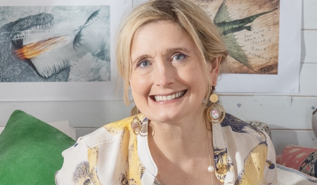 Cressida Cowell smiling with statement earrings on. Siting in front of illustrated dragon artwork on the wall.
