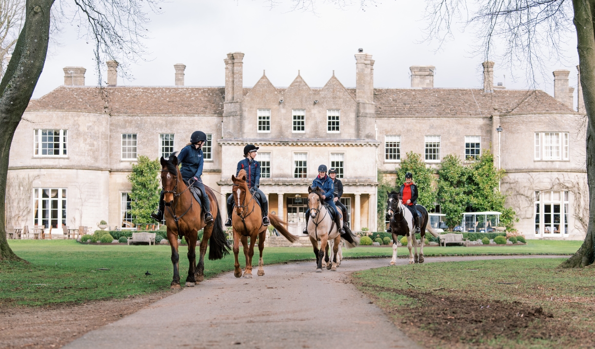 Four horses and riders coming down a path away from a large manor house hotel called Lucknam Park
