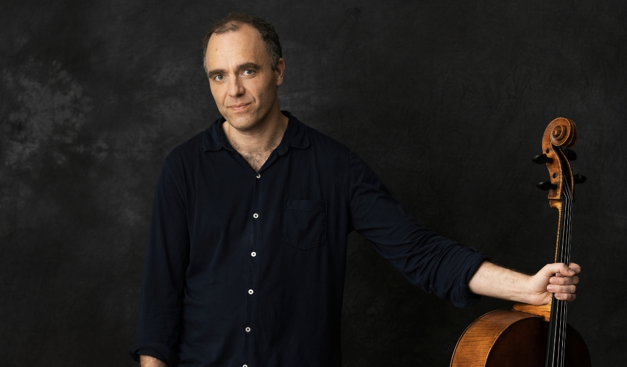 An image of a man holding the neck of a musical instrument in one hand and looking towards the camera.