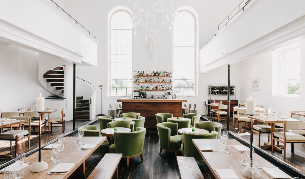 Modern restaurant interior with high ceilings, large arched windows, green cushioned chairs, wooden tables, and a central bar; a spiral staircase is v