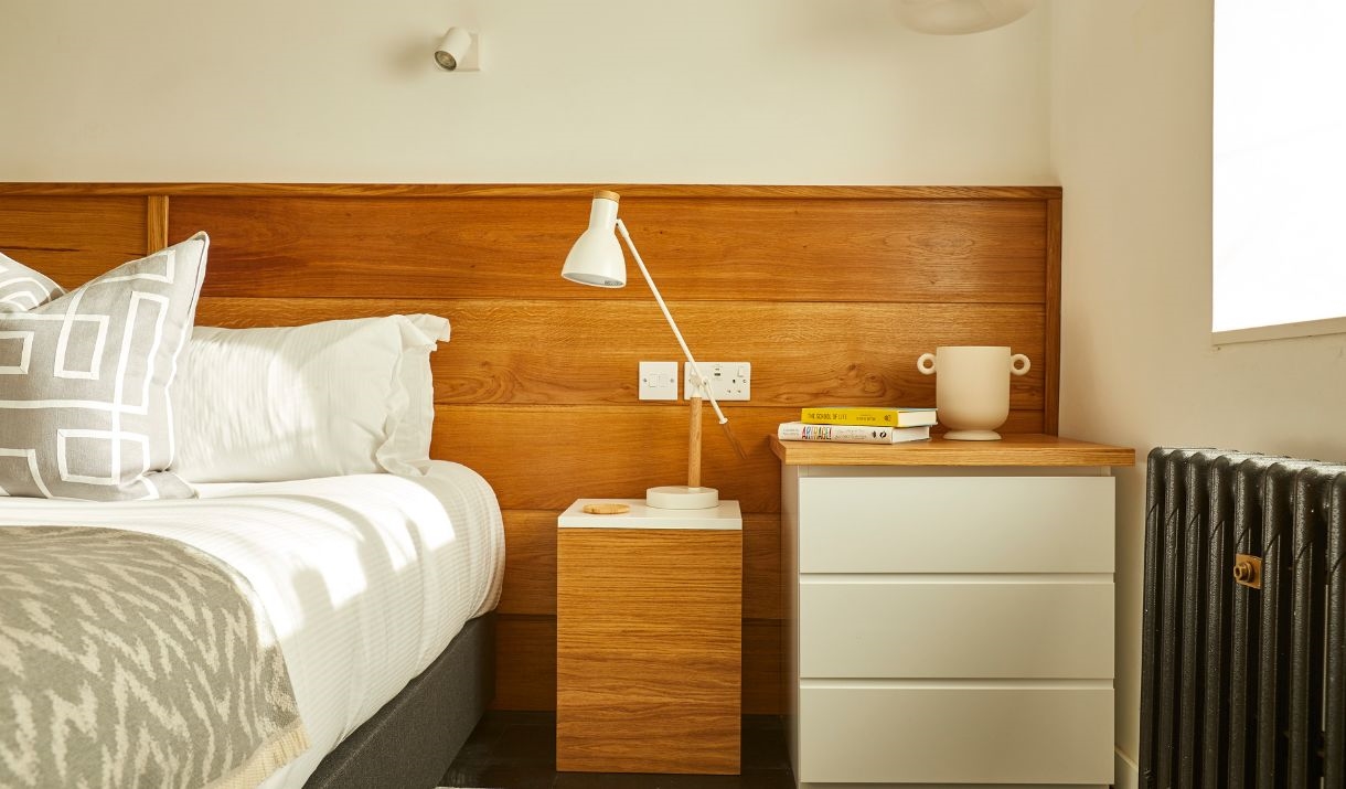 A white bed, gray and white pillows, a wooden headboard, white and wooden nightstands, a white lamp, a beige vase, yellow books, and sunlight streamin