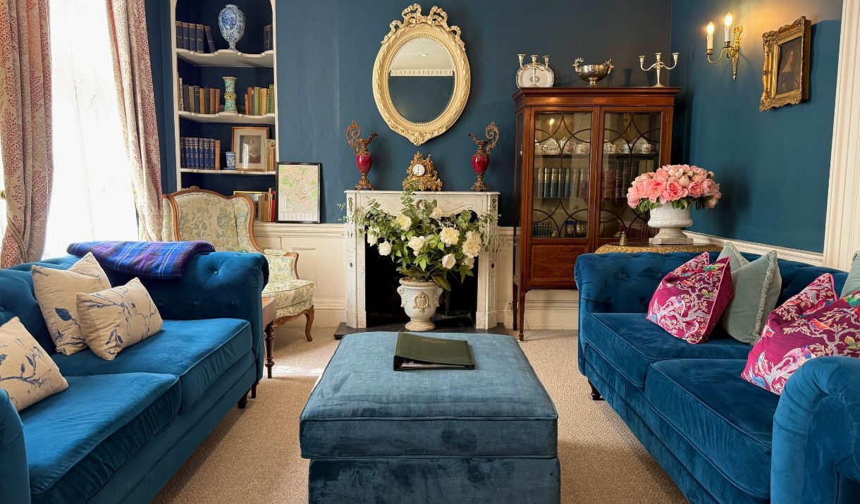 A living room with two blue sofas and an ottoman, a white fireplace and against a blue wall, and an ornate mirror.