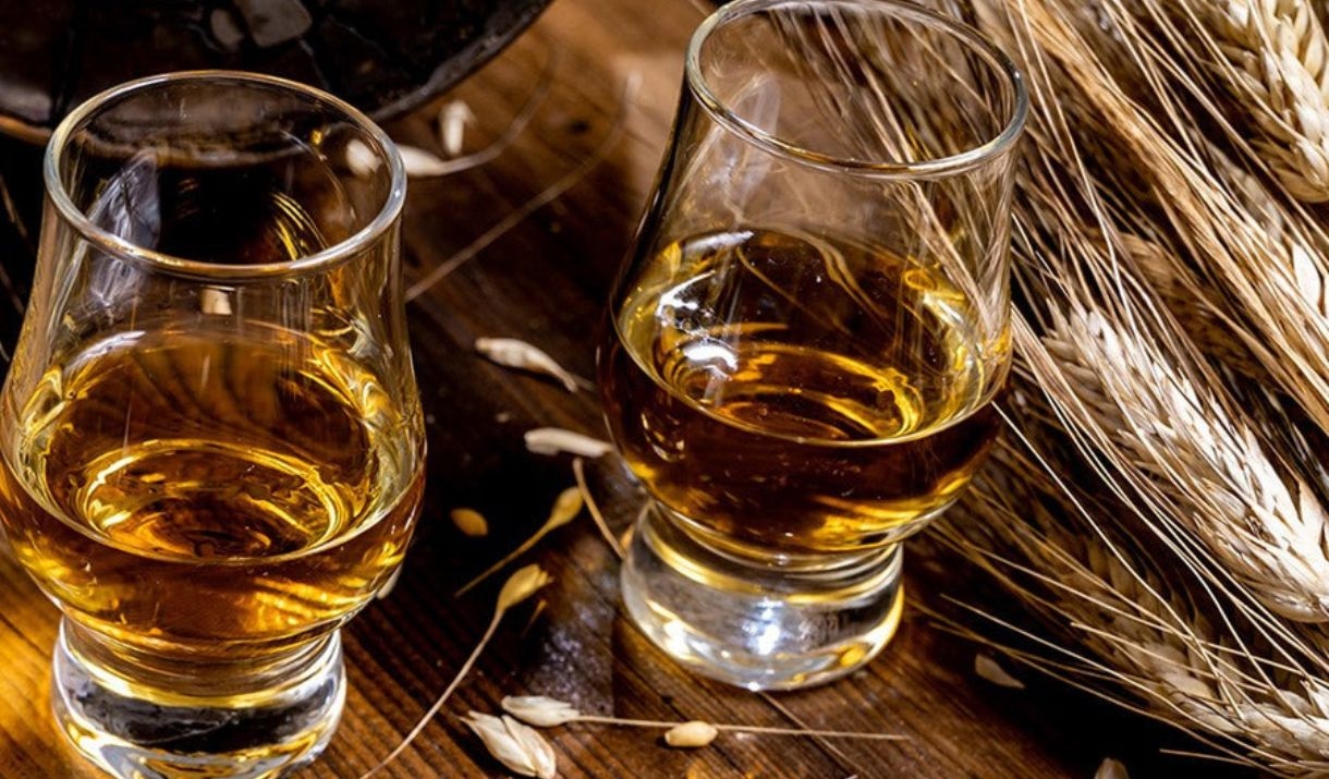 Two clear glass whisky tumblers filled with golden amber liquor, set on a rustic wooden surface with scattered grains and a bundle of wheat stalks nea