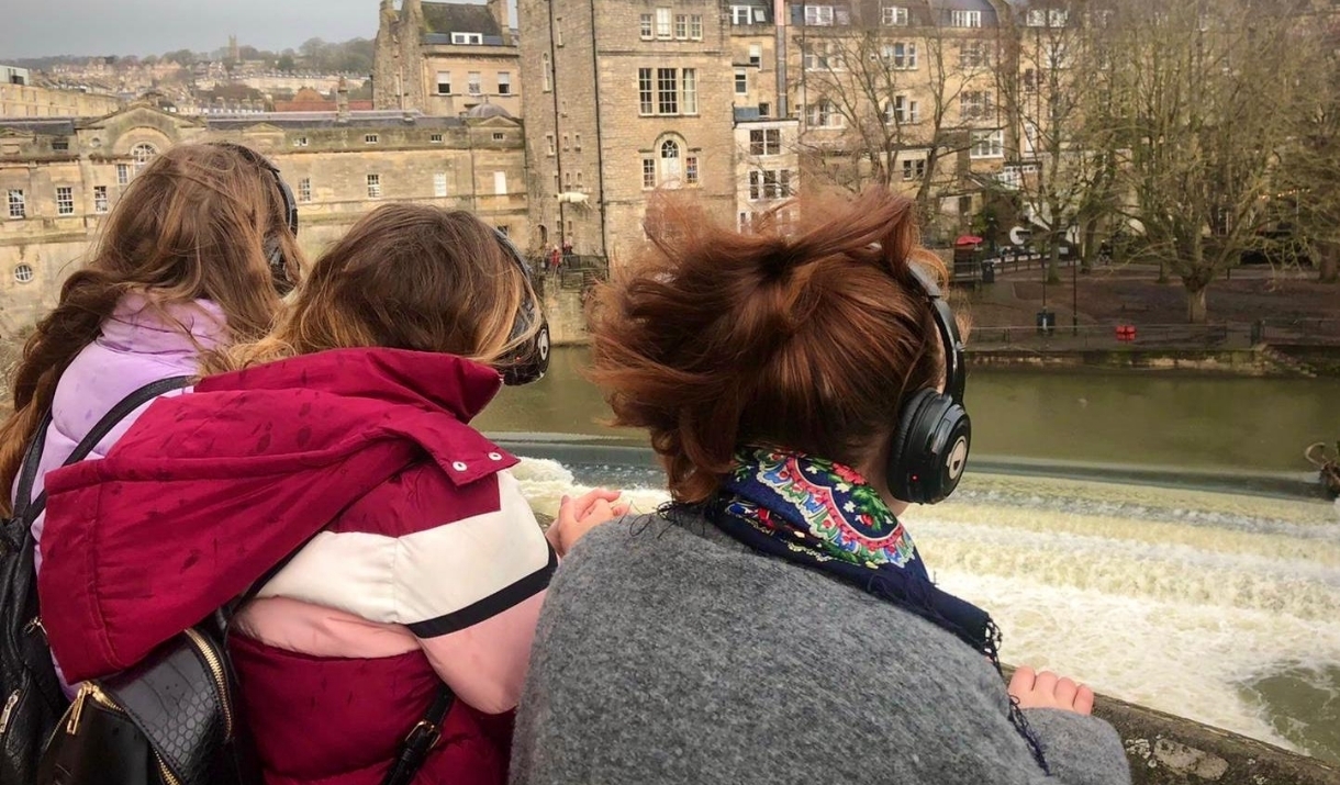 Three people wearing silent disco headphones face away from the camera towards Pulteney Weir in Bath.