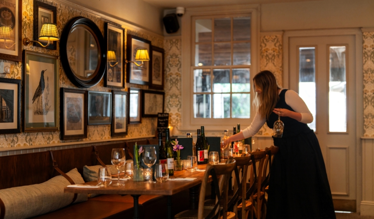 A waitress placing a glass on a long table at The crstal Palace. The table is placed alongside a wall covered in many framed images and a round mirror