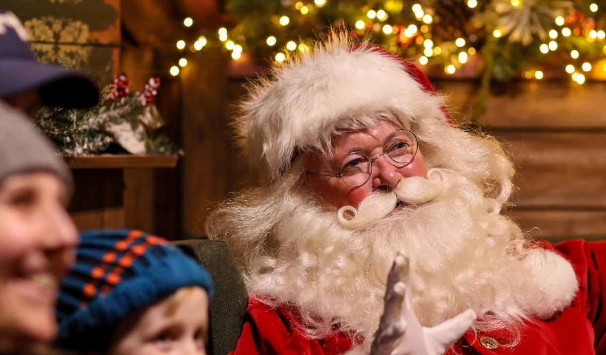 Santa Claus waving warmly at visitors, surrounded by festive holiday lights and decorations.