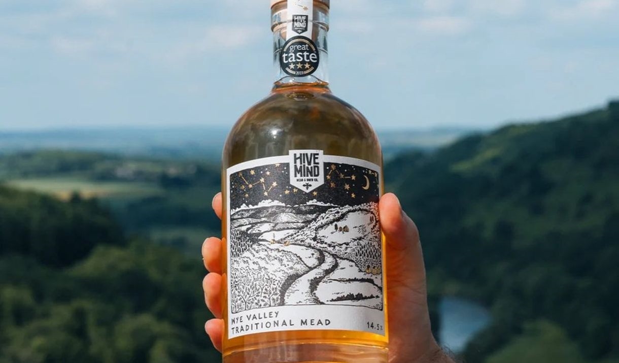 A bottle of bright gold hive mind mead held up in the sun with a green valley behind it.