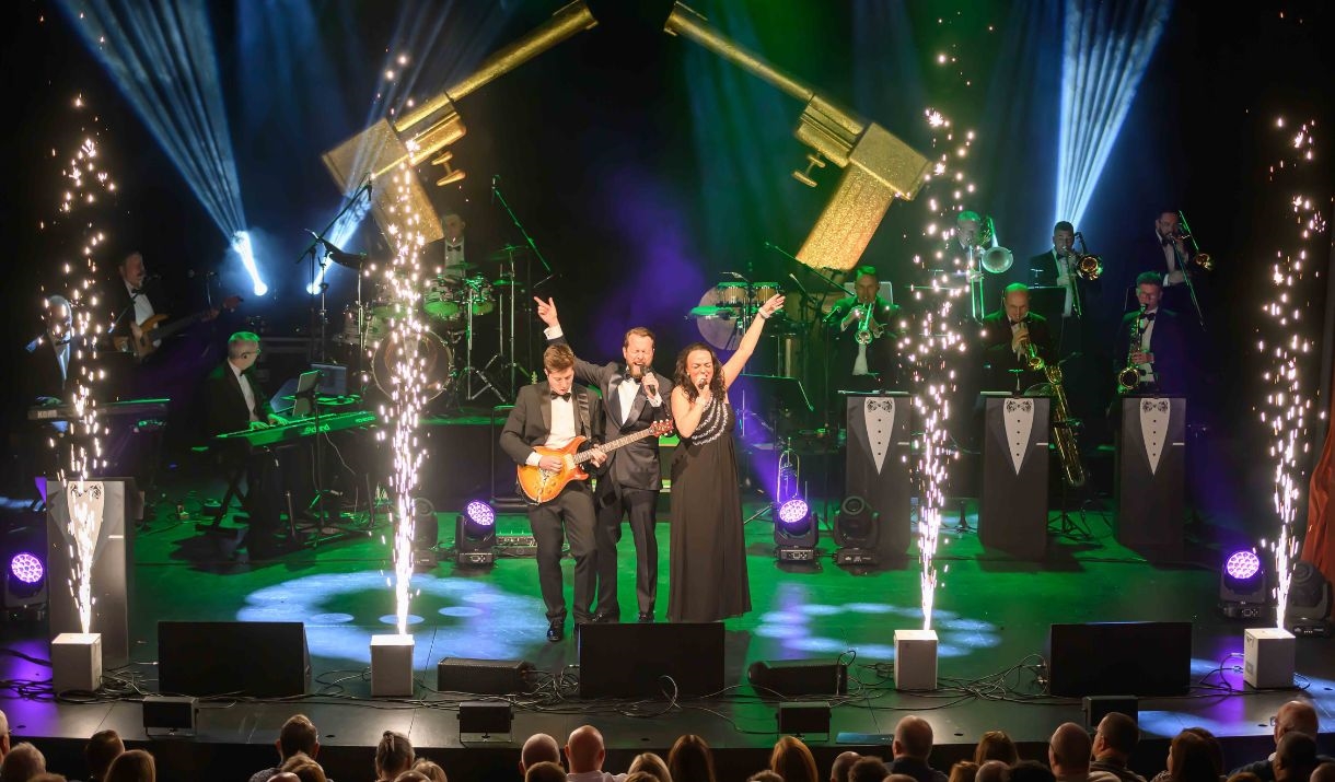 Three singers on stage in black tie dress with pyrotechnics