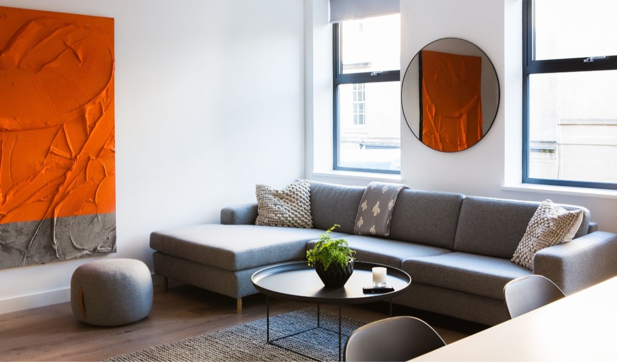 A living room with a large grey corner sofa, round coffee table and a red and grey textured art piece on the wall.