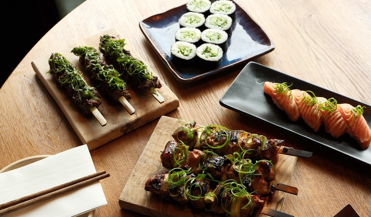 A wooden table with four plates of sushi and meat kebabs on square plates and wooden blocks.