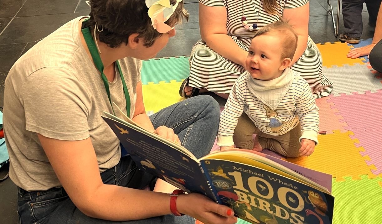 A woman reads a story to a baby sitting on a colourful puzzle floor