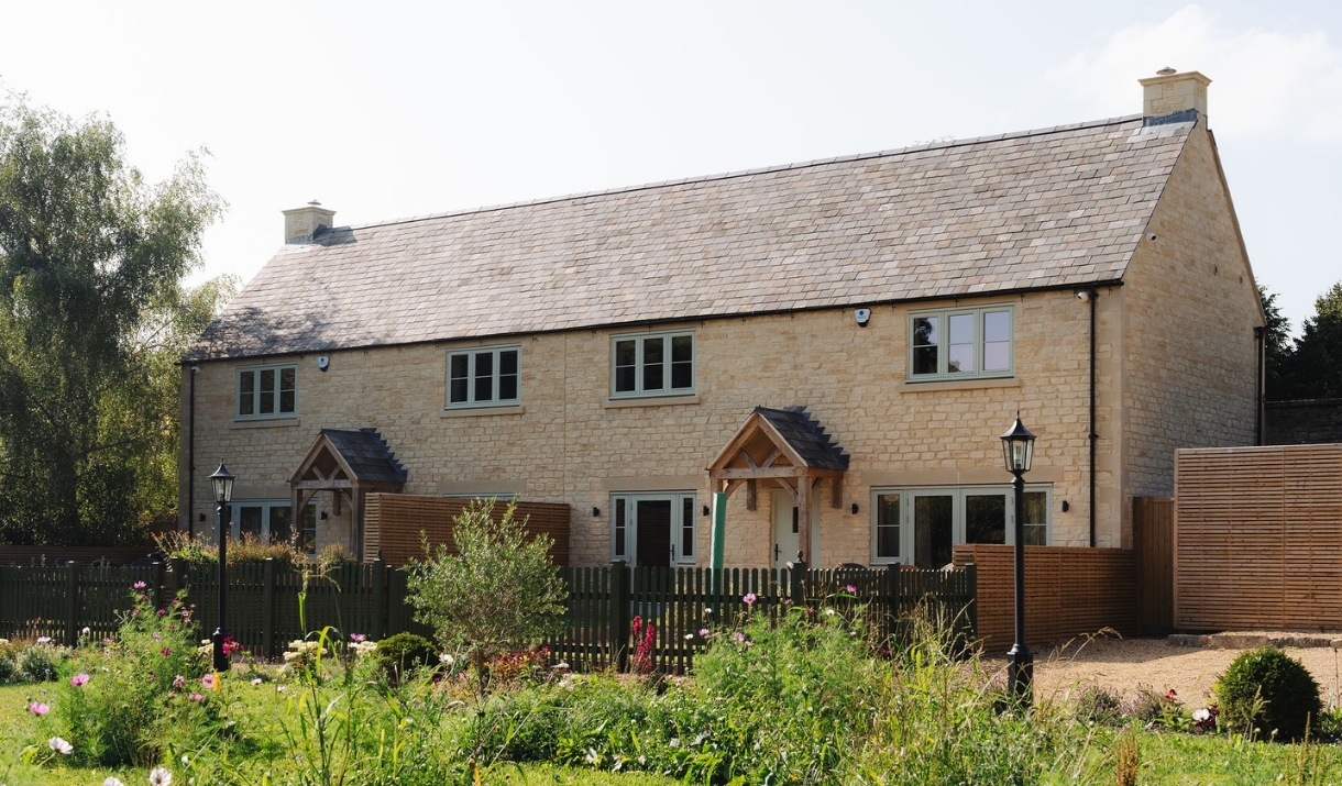 The outside of sandstone row of two cottages with a slate pitch roof and porched front doors with a brown picket fence outside and a garden full of wi