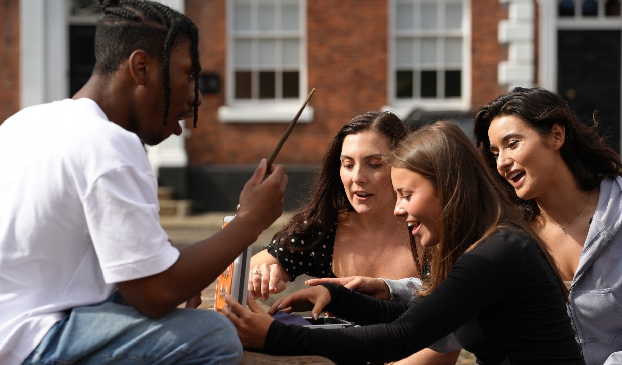 Three women look inside a briefcase while a man looks at a wooden wand with his mouth open.