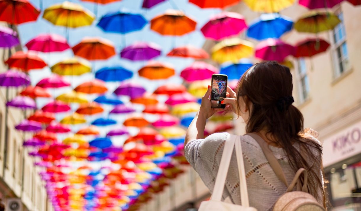 A woman holds a phone up to take a photo of rows of brightly coloured umbrellas hung above the high street