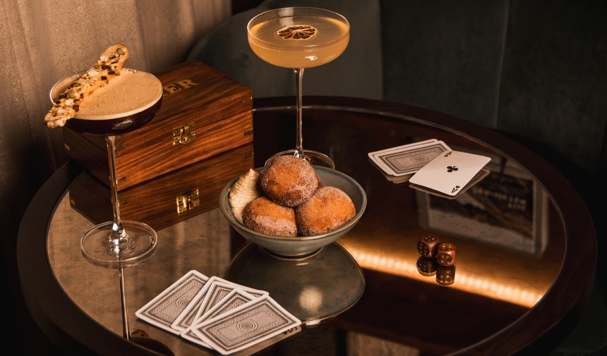 Two cocktails on a table top with a bowl of doughnuts and playing cards.