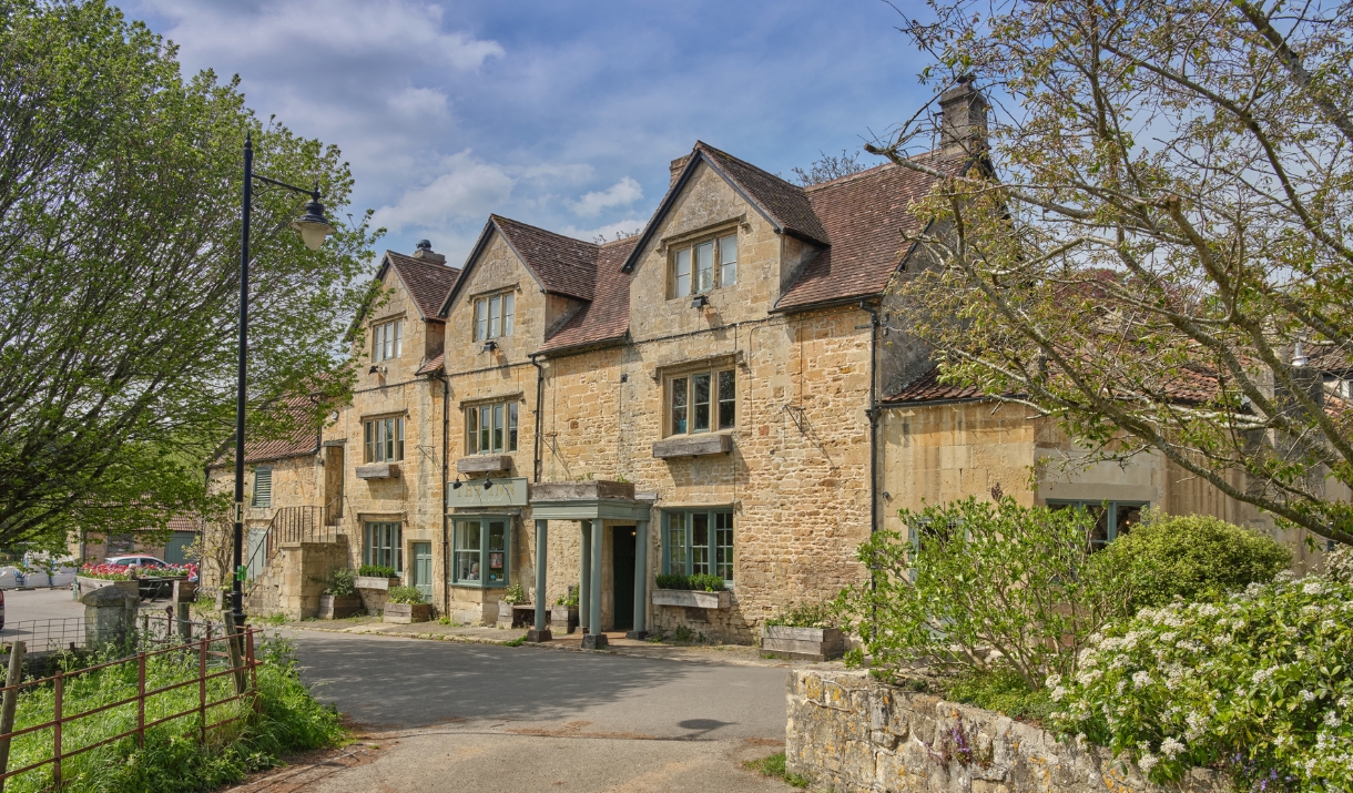A Cotswold stone building on a country road with trees surrounding it.