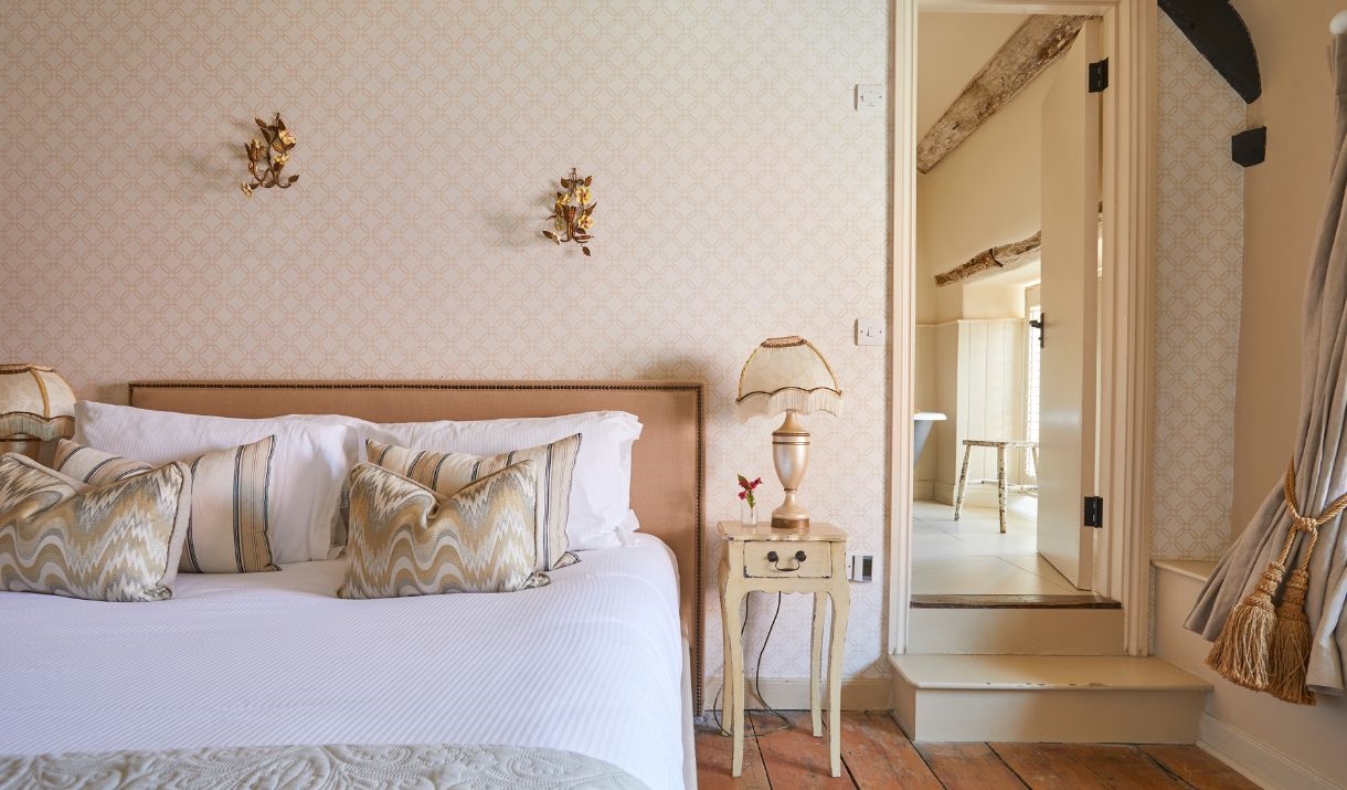 A light coloured bedroom with a bed with white sheets and small bedside table to the right of the bed. A lamp rests atop of the table
