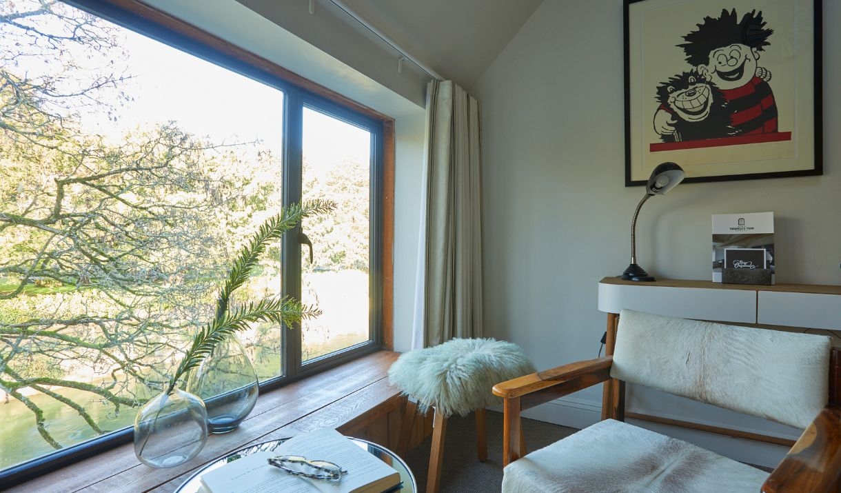 A room with a large chair in the foreground, a stool is in the background and there is a large window to the left of the chair