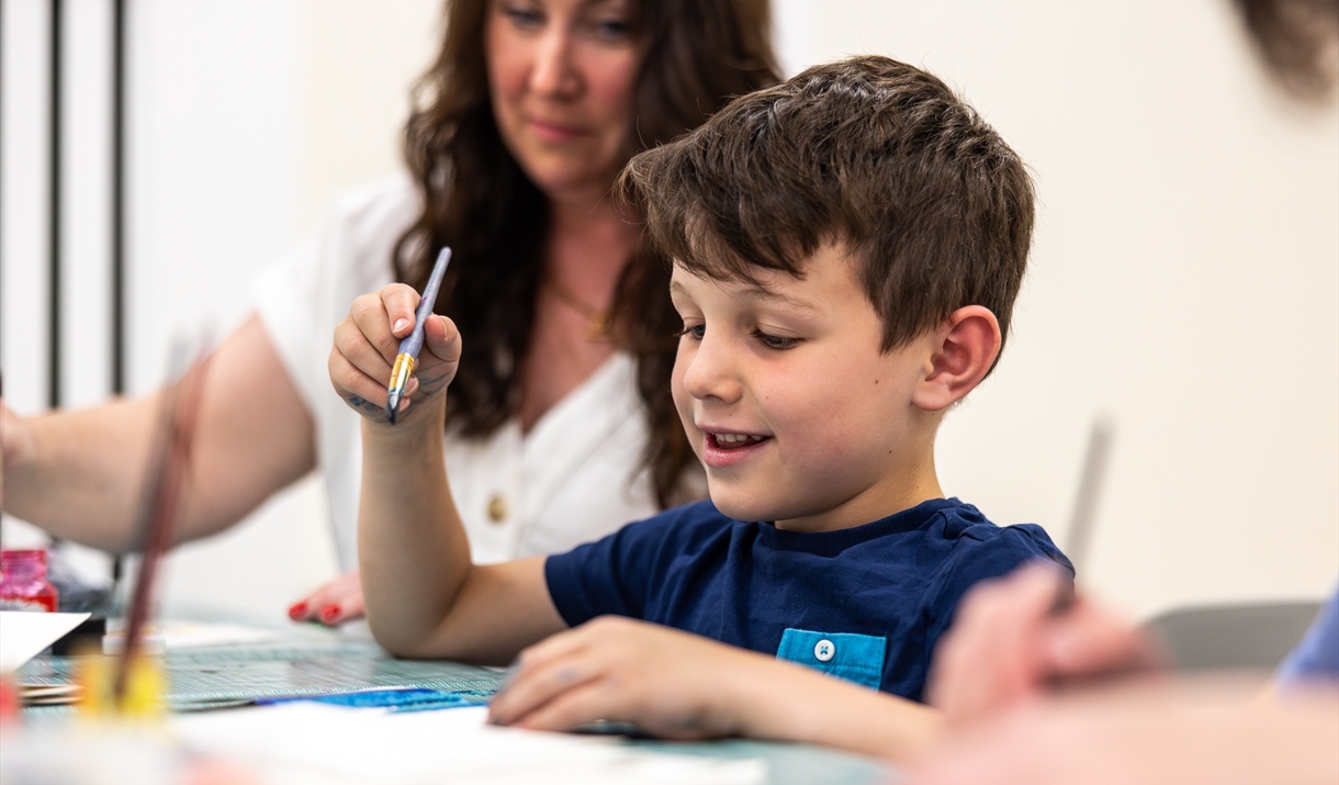 A boy making art with a paintbrush, with his mum looking on.