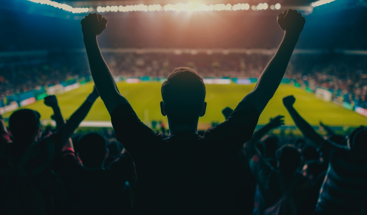 Crowd of football fans celebrating with raised fists at a stadium, floodlights overhead.