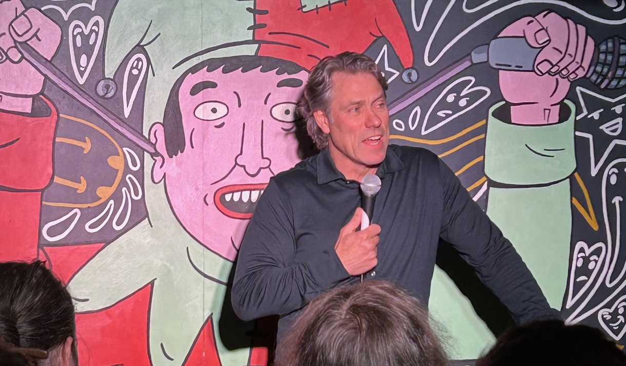 A comedian with a microphone in a black shirt, on stage against a Jester cartoon backdrop