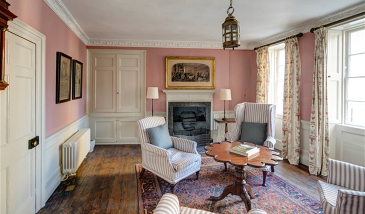 Bright, traditionally furnished snug with pink walls, original wooden floors, a fireplace, striped seating, floral curtains, and a patterned rug, crea