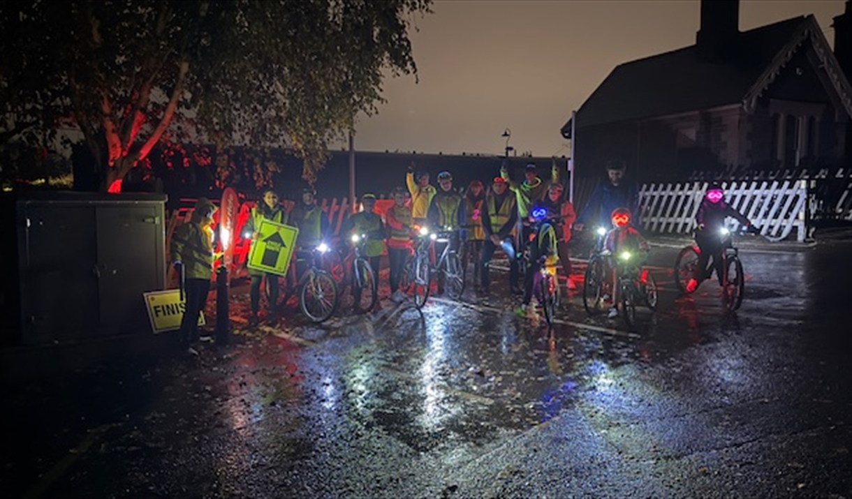 A group of cyclists on bicycles with lights on in the rain outside Bitton Station