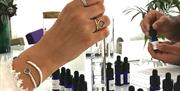 A hand with several rings and bracelets holds a measuring cylinder. Lots of blue fragrance bottles are visible on the table alongside.