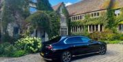 A black luxury car parked on a cobbled road in front of an ivy-covered manor