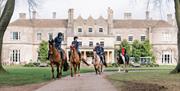Four horses and riders coming down a path away from a large manor house hotel called Lucknam Park