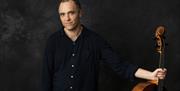 An image of a man holding the neck of a musical instrument in one hand and looking towards the camera.