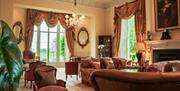 A grand sitting room with large windows and draped curtains, a chesterfield and velvet sofas and chairs around a large marble fireplace with an old oi