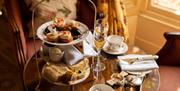 A tiered plate of small cakes and sandwiches along with champagne glasses and tea cups on a small round mahogany table
